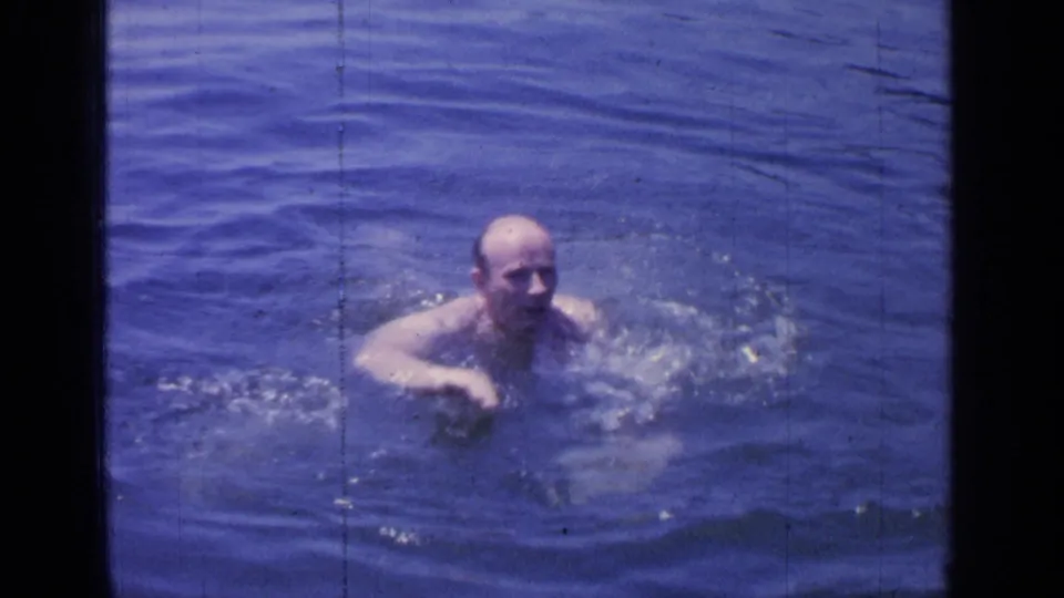 1964 Greenwood Lake frame of a bald adult swimmer approaching through rippled blue water.