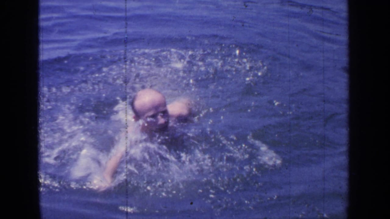 At Greenwood Lake in 1964, an adult man swims toward the camera through rippled blue water in grainy 8mm footage.