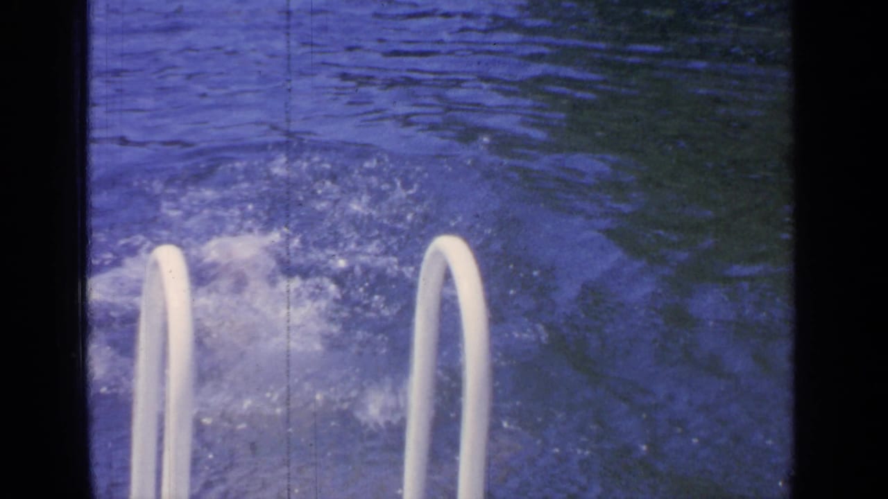 1964 Greenwood Lake home-movie frame showing splashing pool water beside white ladder rails with blue 8mm color and film scratches.