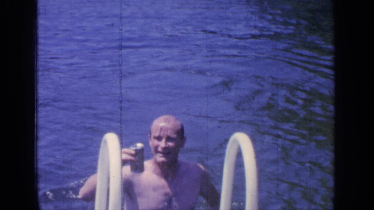 Shirtless adult man smiles in pool water while holding a can near ladder rails.