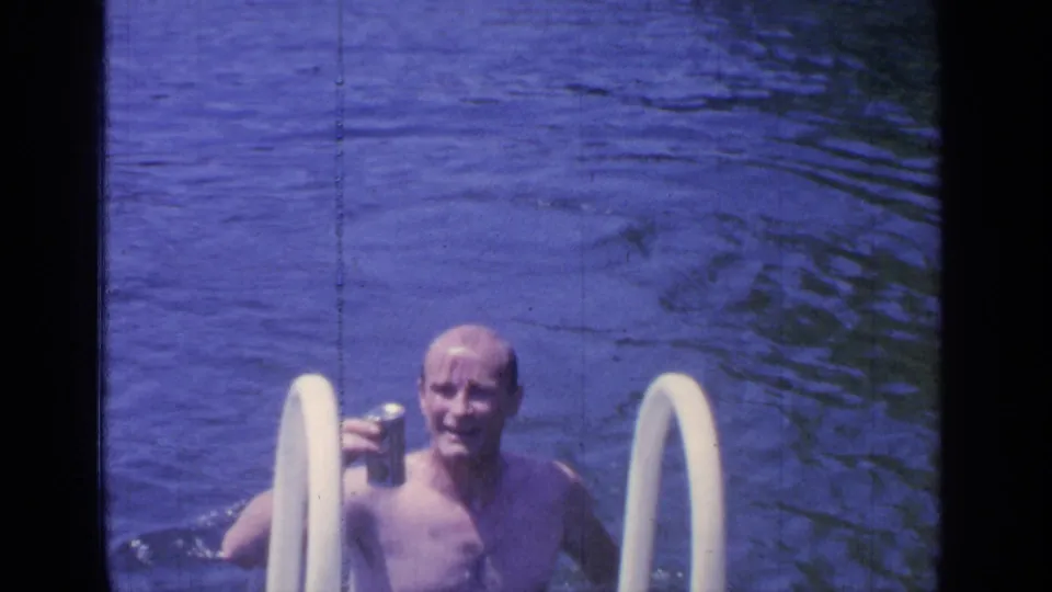 1964 Greenwood Lake home-movie frame of a shirtless adult man smiling in pool water while holding a small can beside white ladder rails.
