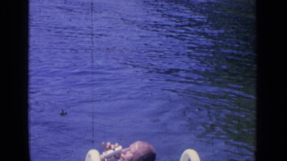 1964 Greenwood Lake home-movie frame showing the man partly cropped as he drinks from a can in blue-toned pool water.