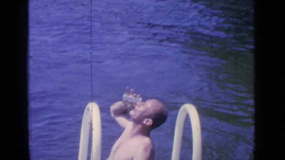 1964 Greenwood Lake home-movie frame of a shirtless adult man drinking from a can while standing or floating near the pool ladder.