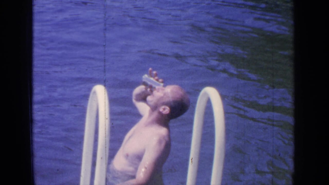Man standing in lake water beside a white ladder while drinking from a can.