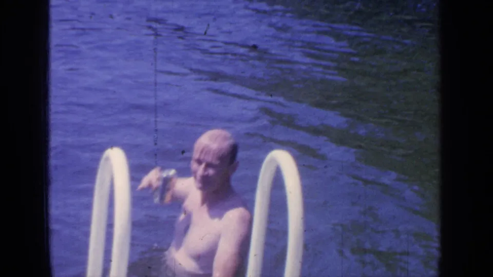 The man lowers the can while standing in Greenwood Lake water near the white ladder, with his face visible toward the camera.