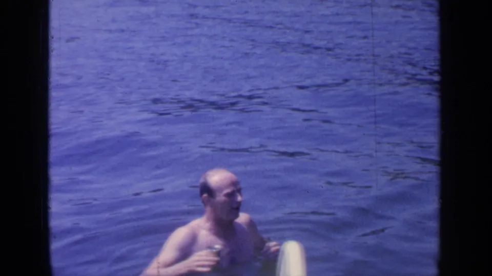 In the 1964 Greenwood Lake footage, the man remains chest-deep in rippled water with the can in hand and the ladder partly visible.