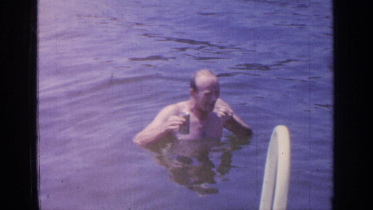 At Greenwood Lake in 1964, a shirtless man stands chest-deep in water holding a beer can beside a white swim ladder.