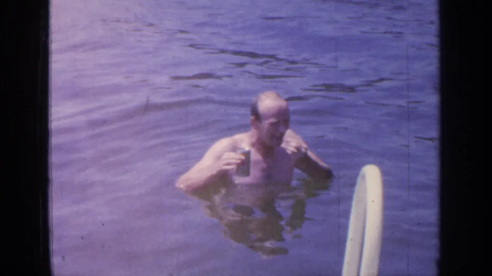 At Greenwood Lake in 1964, a shirtless man stands chest-deep in water holding a beer can beside a white swim ladder.