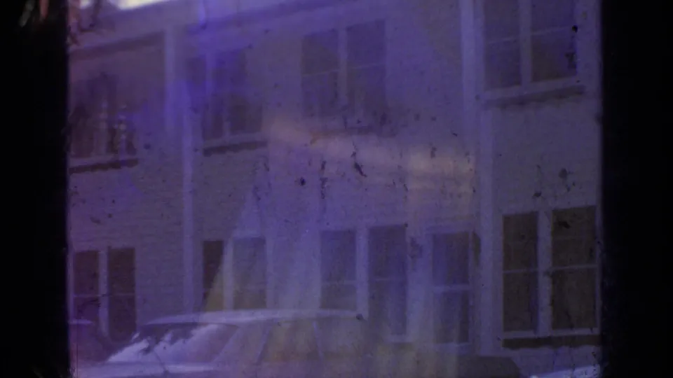 A blue-purple home-movie frame shows a pale building facade with windows and parked cars near the lakeside setting.