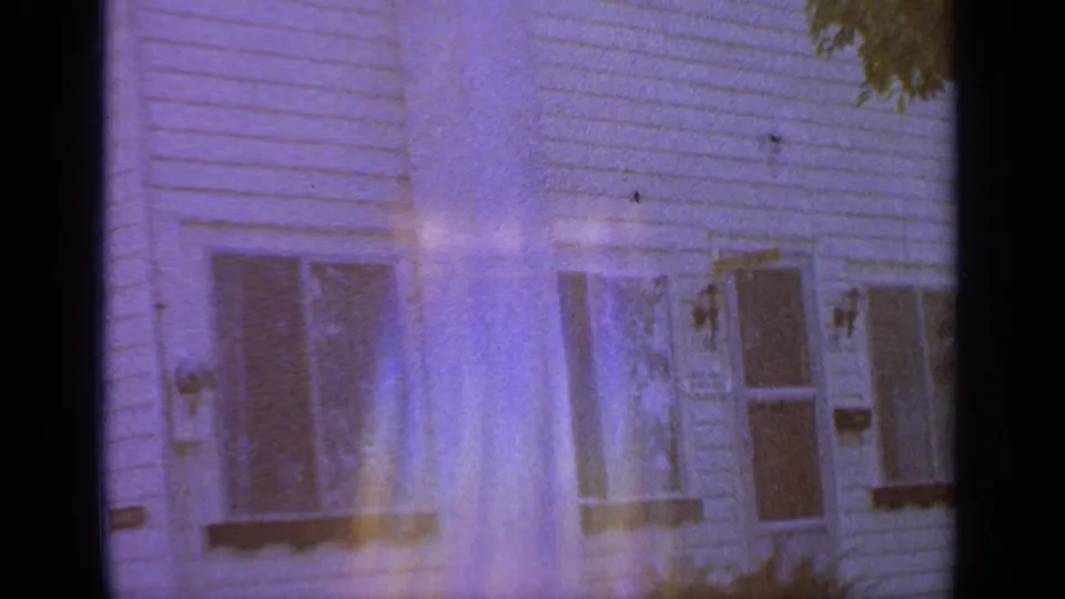 1964 home-movie frame showing a white clapboard wall with lower windows, shutters, foliage, and soft purple flare.