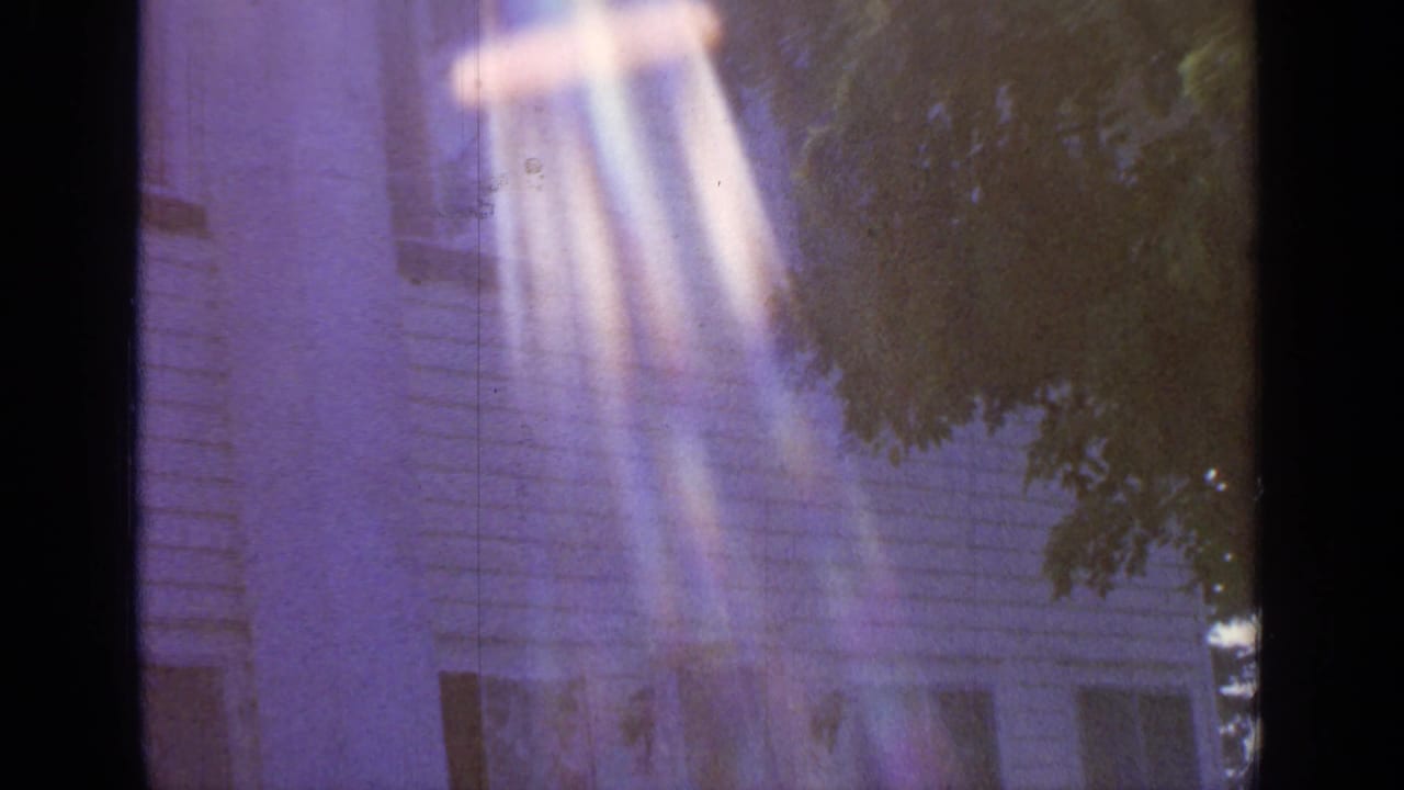1964 8mm frame from Greenwood Lake showing the upper portion of a white clapboard house behind leafy branches with purple color cast and bright vertical flare.