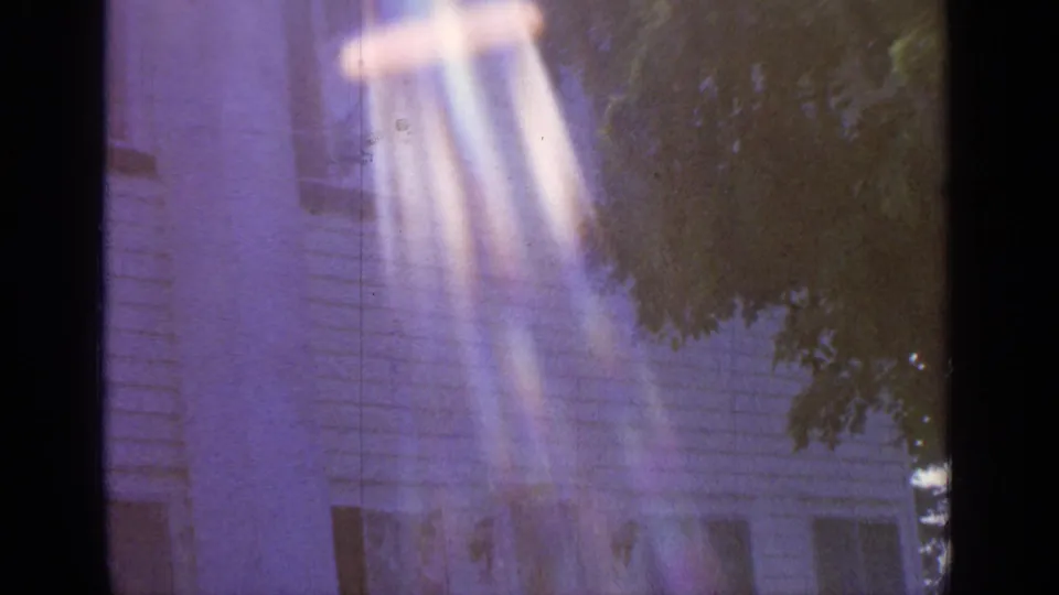 1964 8mm frame from Greenwood Lake showing the upper portion of a white clapboard house behind leafy branches with purple color cast and bright vertical flare.