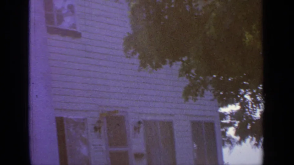 Greenwood Lake house exterior in a purple-tinted 8mm frame, with clapboard siding, shuttered windows, and dark tree foliage crossing the upper right.