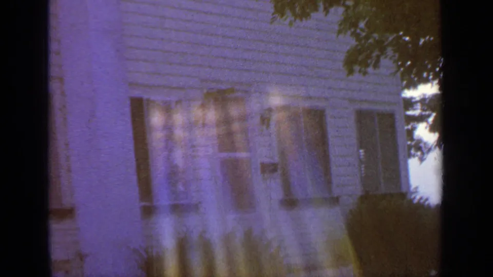 8mm frame of the lower clapboard house exterior in Greenwood Lake, with shuttered windows, shrubs near the wall, and translucent lens flare over the siding.
