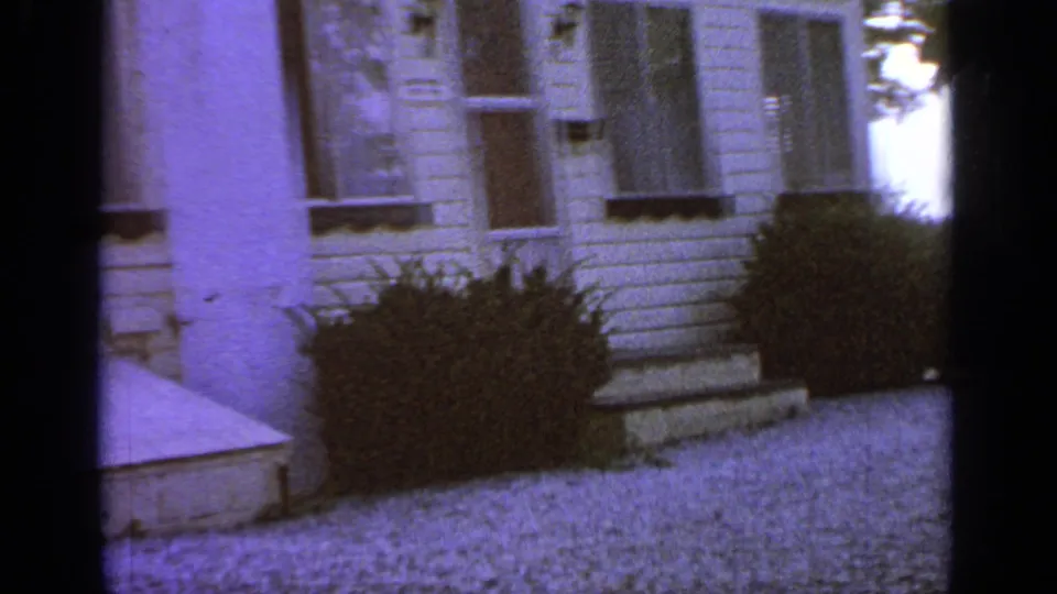 1964 Greenwood Lake home-movie frame showing the front entry area of a white clapboard house, including the door, porch steps, bushes, and pale ground in front.