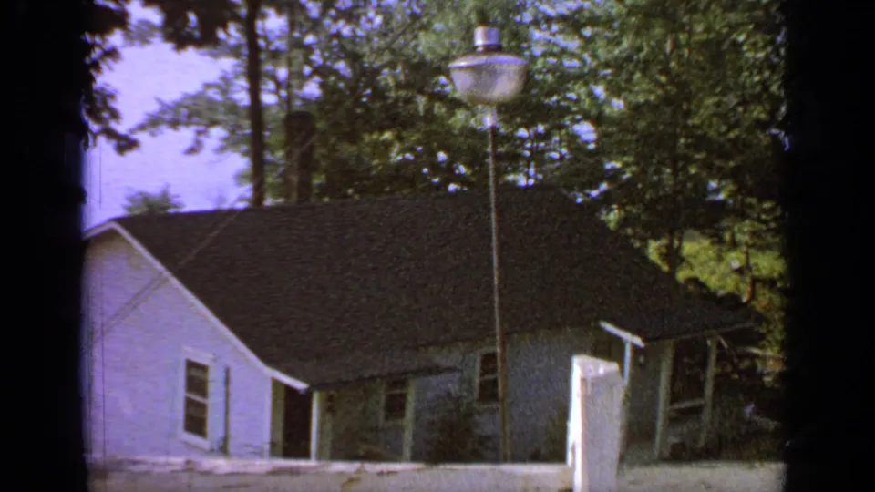 1964 Greenwood Lake frame with a small white building, dark roof, surrounding trees, gravel yard, and a pole-mounted lamp.
