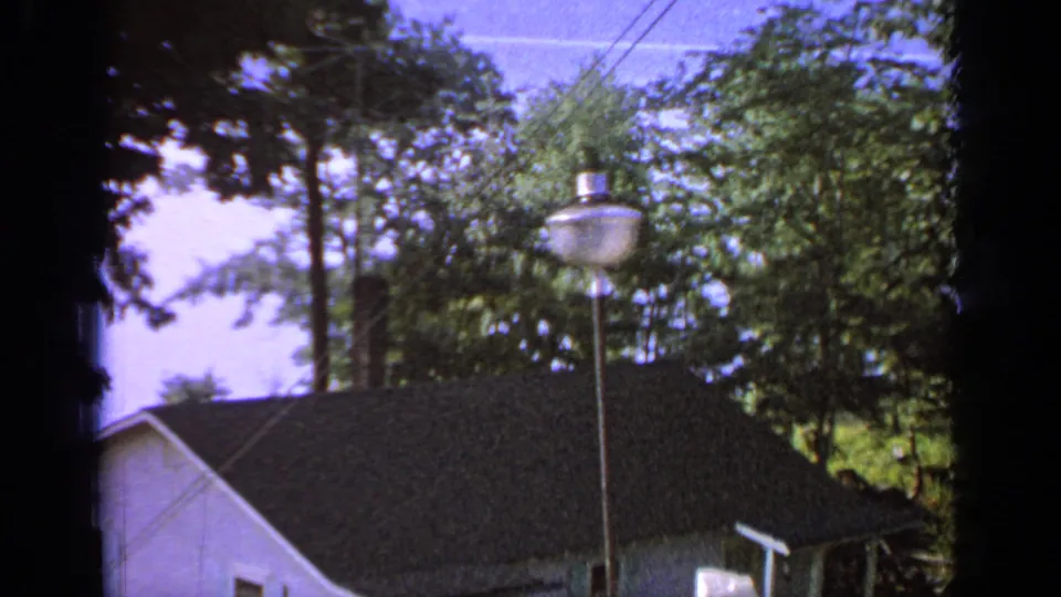 Greenwood Lake home-movie frame showing a white lakeside building, dark roof, tall yard lamp, trees, overhead wires, and sky.