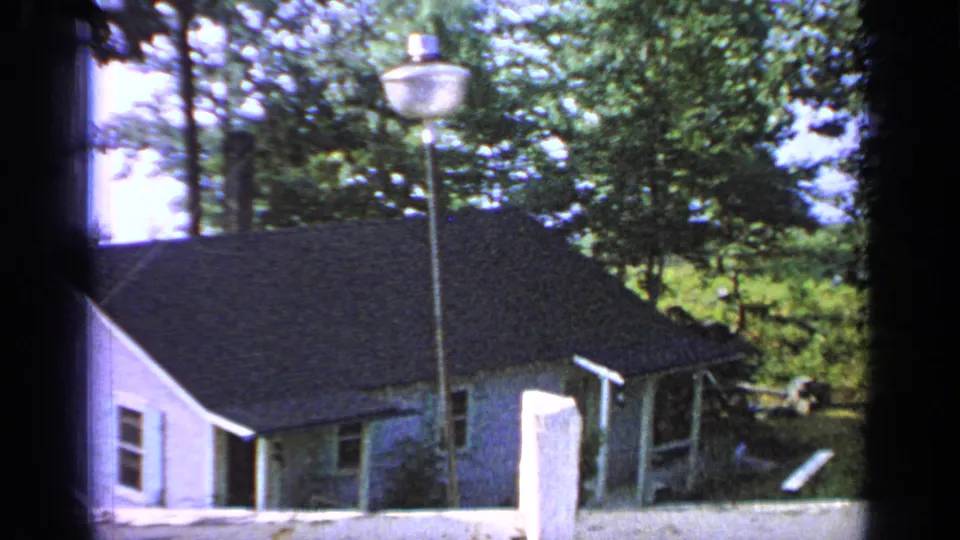 1964 Greenwood Lake home-movie frame showing a white house, dark roof, street lamp, low wall, and green trees.