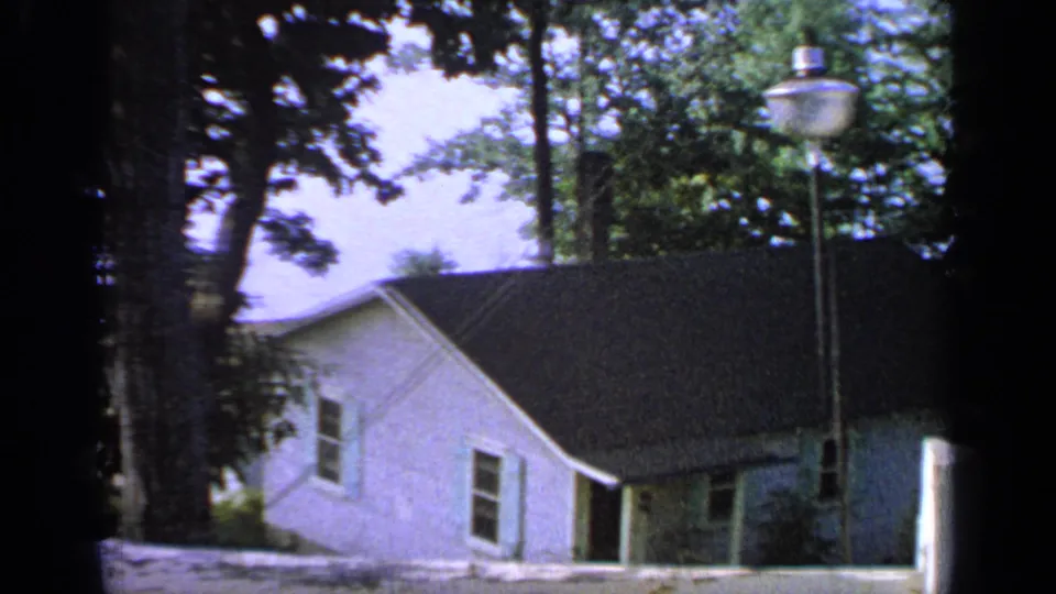 1964 Greenwood Lake frame of a white residential house, dark roof, shaded trees, and a foreground street lamp.
