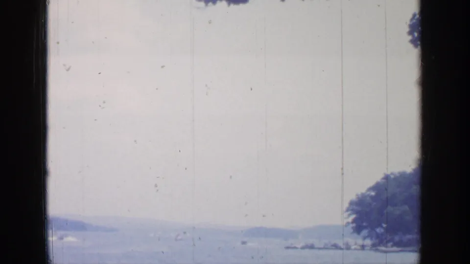 Window-framed view of Greenwood Lake with hazy water, distant hills, shoreline trees, and small boats in the distance.