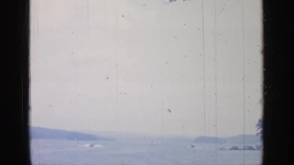 1964 Greenwood Lake view across pale water toward low hills, with small boats faintly visible and heavy 8mm film wear.