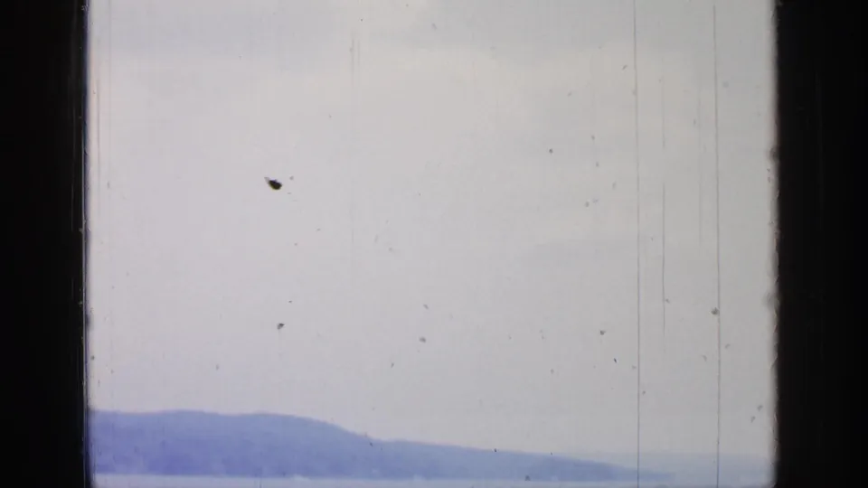 Overexposed 8mm frame of Greenwood Lake and low hills, framed by a dark window edge and faint shoreline detail.