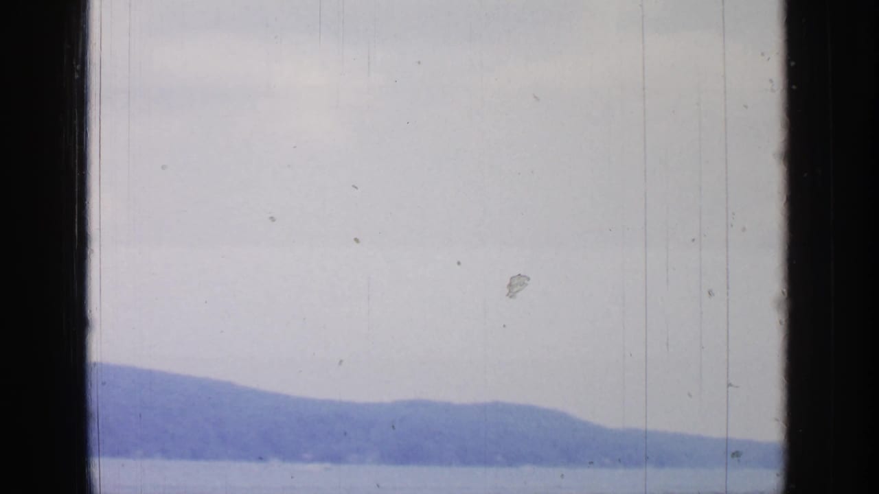 1964 Greenwood Lake footage showing calm water, distant wooded hills, pale sky, and dark vertical framing in soft color 8mm.
