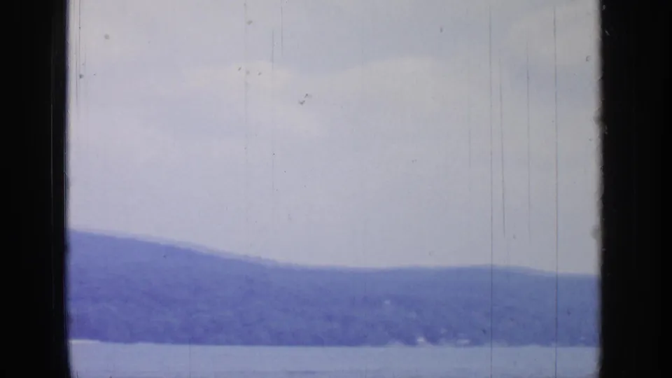 A soft 8mm view of Greenwood Lake shows a low tree-covered ridge, calm foreground water, bright sky, and heavy dark framing at the sides.