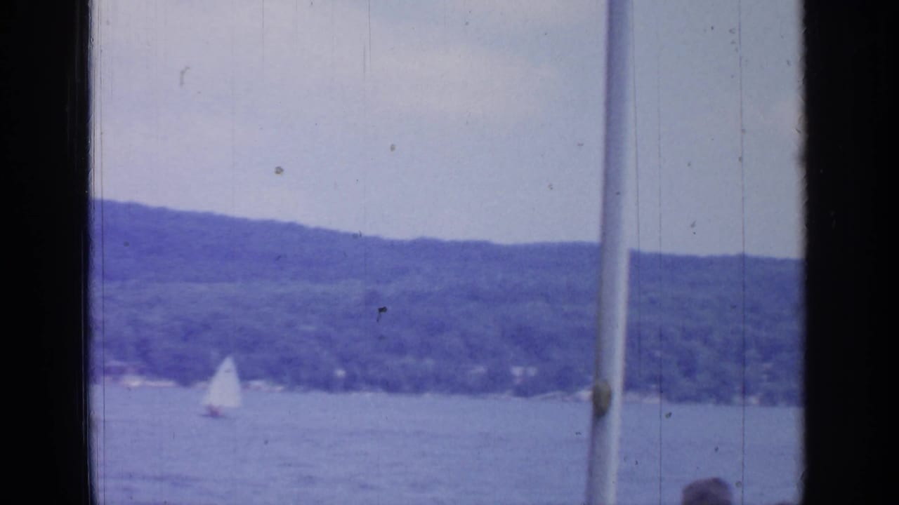 Small white sailboat on a lake below wooded hills.