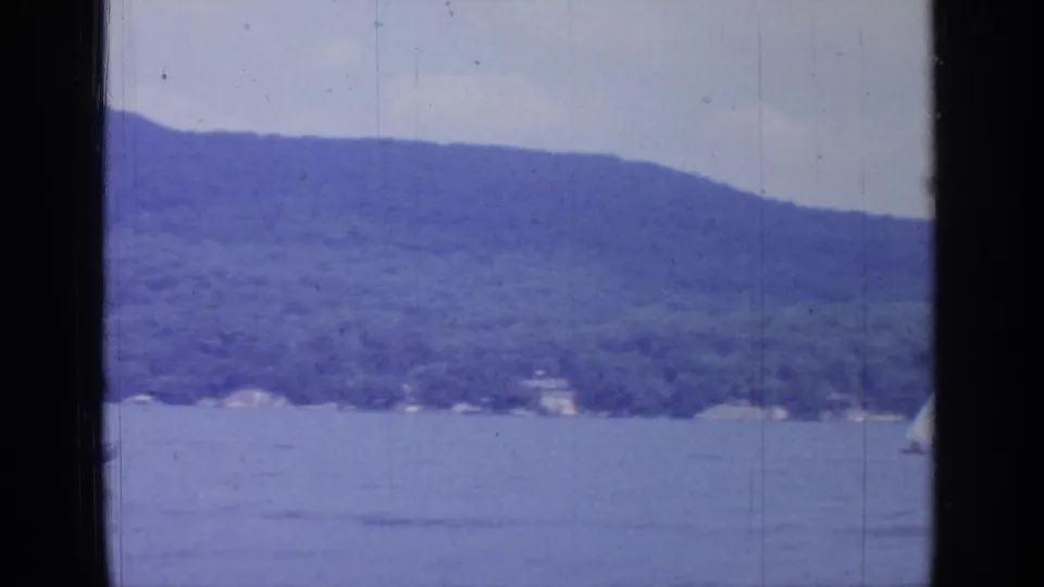 Wide Greenwood Lake view with wooded hills, blue water, and a partial edge of the filming boat in 1964 8mm footage.