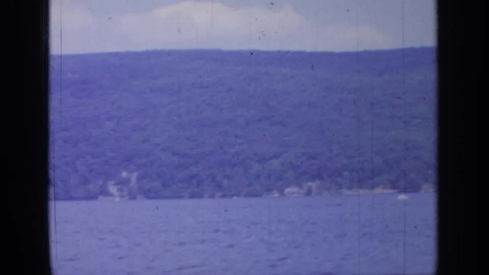 1964 Greenwood Lake view from a moving vessel, showing blue water, distant wooded hillside, pale sky, and dark window framing.