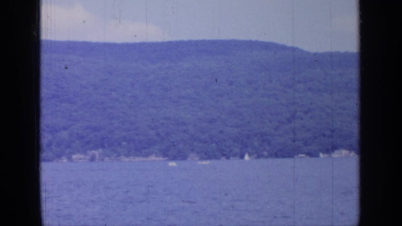 Greenwood Lake shoreline in 1964, with blue water, tree-covered hills, faint buildings, and small white sail shapes in the distance.