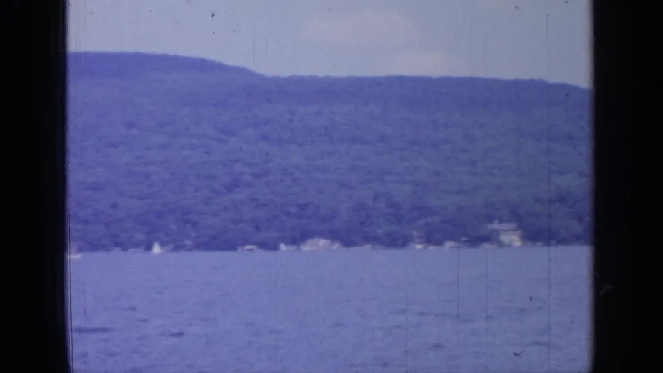 Vintage 8mm view of Greenwood Lake, with open water, hazy wooded hills, shoreline houses or structures, and a small white sail shape.