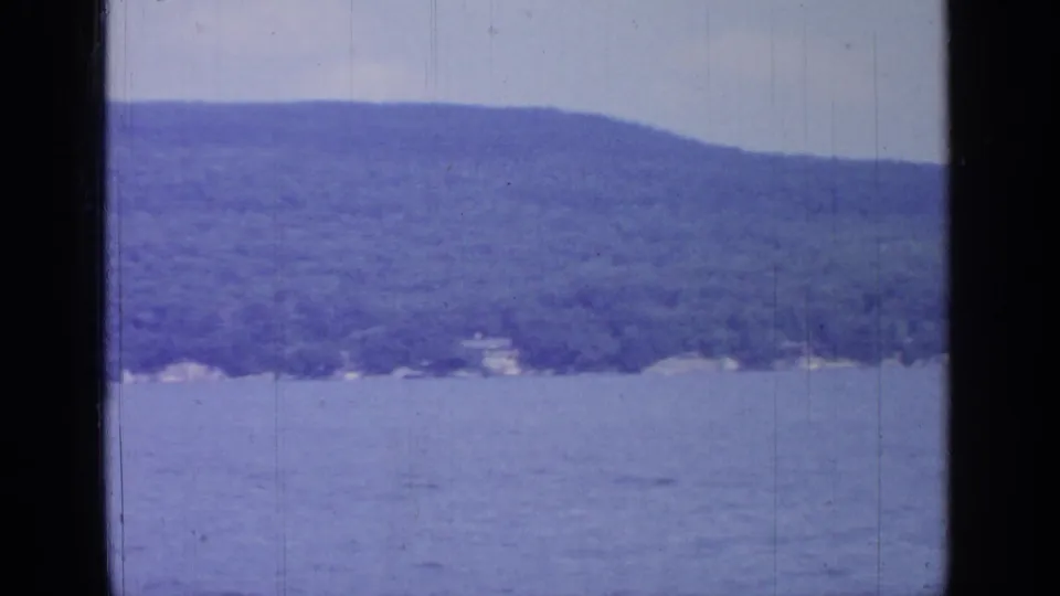1964 Greenwood Lake scenery from a vessel, showing calm blue water, a wooded hillside, faint shoreline buildings, and aged film texture.