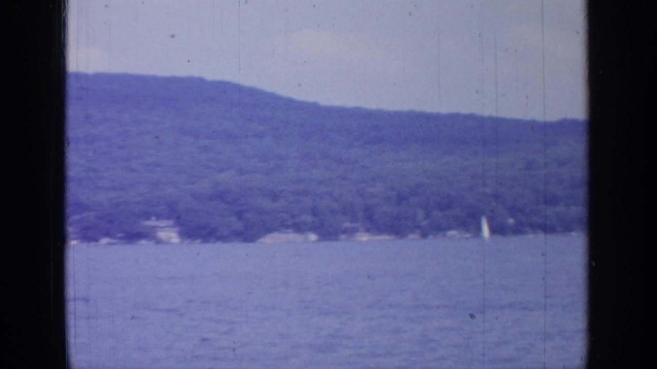 1964 8mm view of Greenwood Lake with calm blue water, tree-covered hills, pale sky, and dark frame edges.