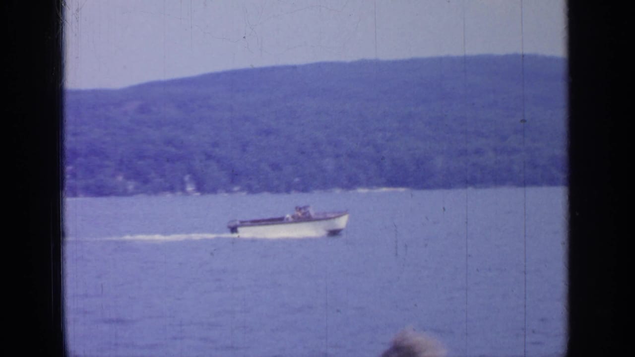 Small motorboat crosses the lake below wooded hills, leaving a white wake.