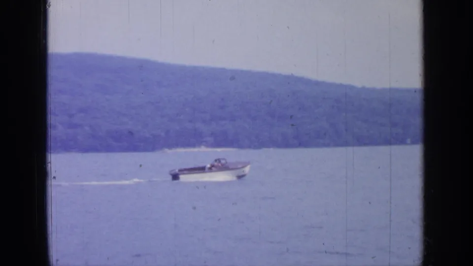 Small motorboat crossing Greenwood Lake with a low white wake, distant wooded shoreline, and soft 1960s 8mm color.