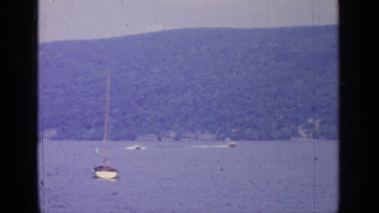 1964 Greenwood Lake frame with an anchored sailboat in the foreground and distant boats near a wooded shoreline.