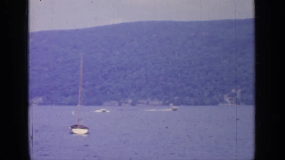 1964 Greenwood Lake frame with an anchored sailboat in the foreground and distant boats near a wooded shoreline.
