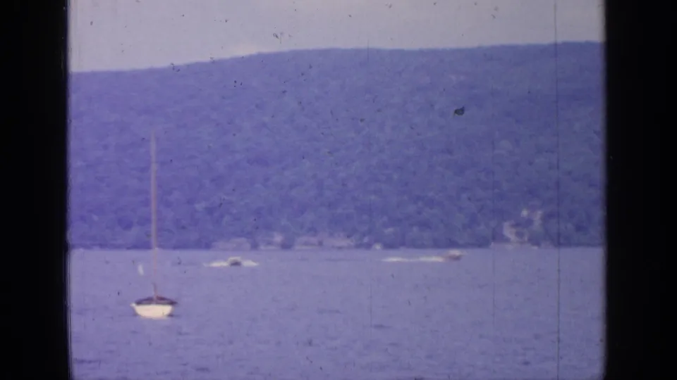 1964 Greenwood Lake 8mm view with a sailboat near the foreground and small boats along the distant shore.