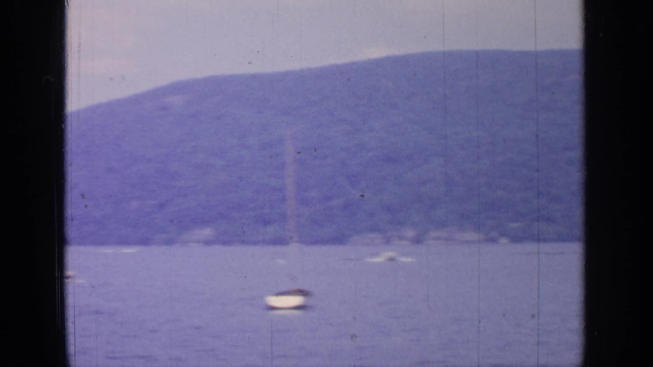 1964 Greenwood Lake view with a small sailboat on calm water below wooded hills and a hazy sky.