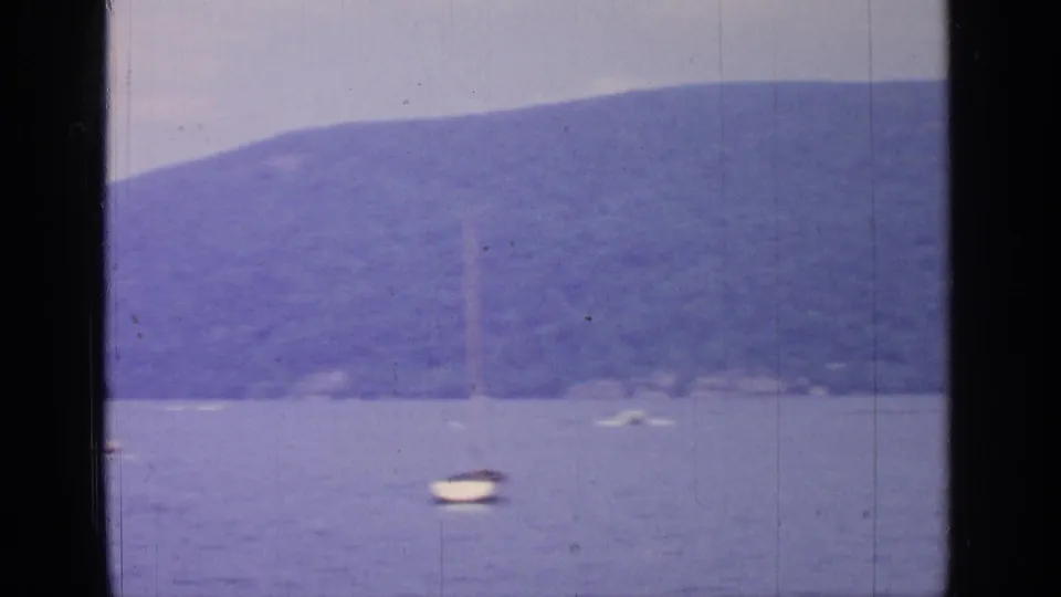 1964 Greenwood Lake view with a small sailboat on calm water below wooded hills and a hazy sky.