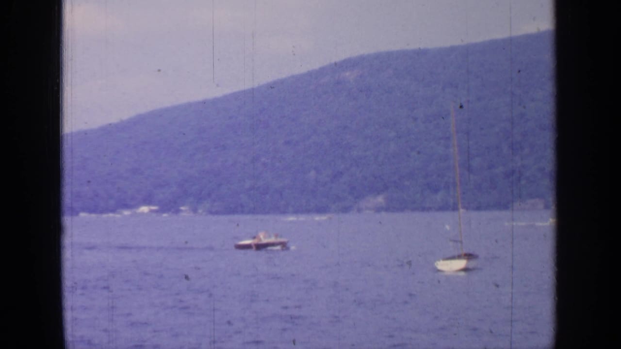 Motorboat and sailboat share the water at Greenwood Lake, with wooded hills and soft 8mm color in the background.