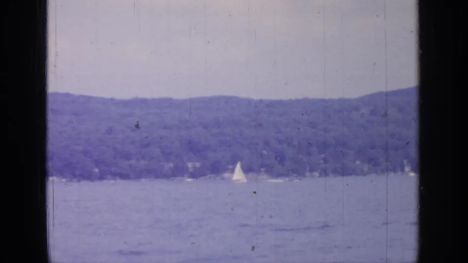 Distant sailboats move across Greenwood Lake against low forested hills in soft, faded 8mm footage.