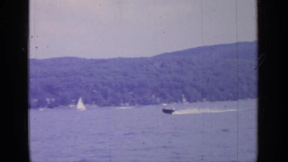 A motorboat leaves a bright wake across Greenwood Lake while sailboats remain farther off near the wooded shore.