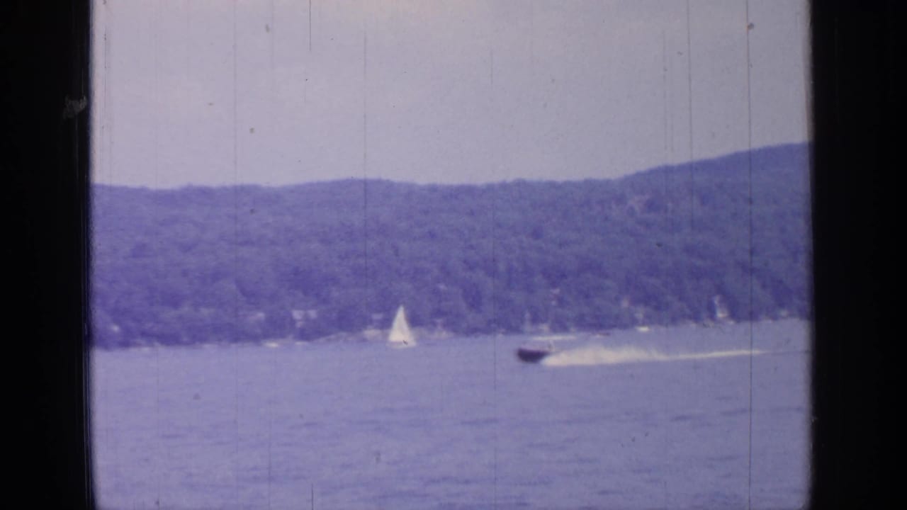 Sailboat and speedboat on a lake with wooded hills in the distance.