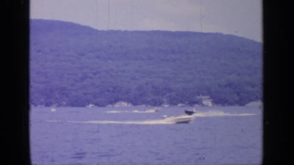 1964 Greenwood Lake home-movie frame showing a speedboat wake near distant lakeside houses and wooded hills.
