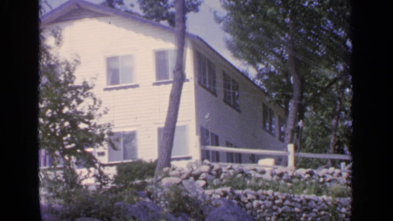 1964 Greenwood Lake cottage exterior with white siding, porch rail, trees, and a stone retaining wall in a wooded setting.