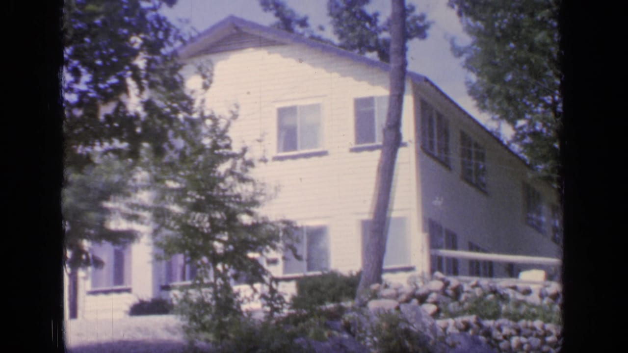 White cottage at Greenwood Lake, New York, viewed through trees with windows, siding, porch rail, and stone landscaping visible.