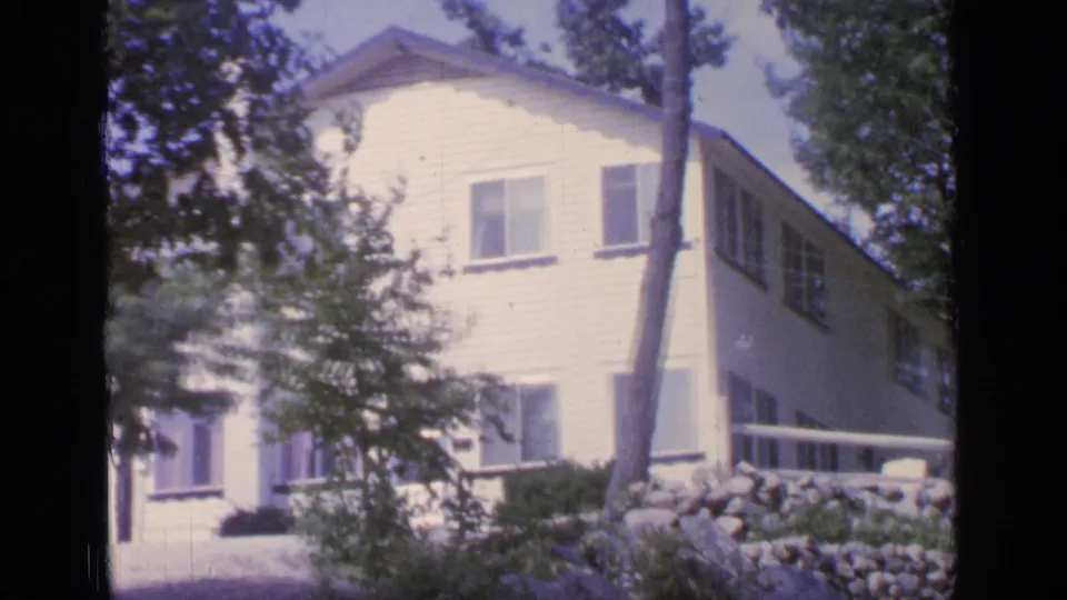 White cottage at Greenwood Lake, New York, viewed through trees with windows, siding, porch rail, and stone landscaping visible.